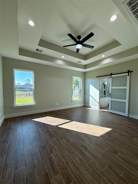 This bright room features a tray ceiling with recessed lighting and a modern ceiling fan. It has wood-look tile flooring, two large windows for natural light, and a sliding barn door leading to an adjoining space. This bright room features a tray ceiling with recessed lighting and a modern ceiling fan. It has wood-look tile flooring, two large windows for natural light, and a sliding barn door leading to an adjoining space.