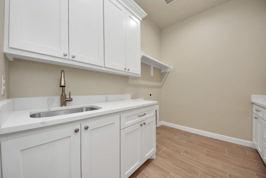 Here is the laundry room with the same white custom cabinetry, a sleek stainless-steel sink, and wood look tile flooring, offering ample storage and workspace.