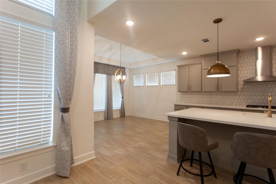 A bright breakfast nook flows seamlessly from the kitchen, framed by oversized windows, custom drapery, and a designer chandelier. The kitchen showcases a gas cooktop, stainless vent hood, white subway tile, and a built-in breakfast bar.