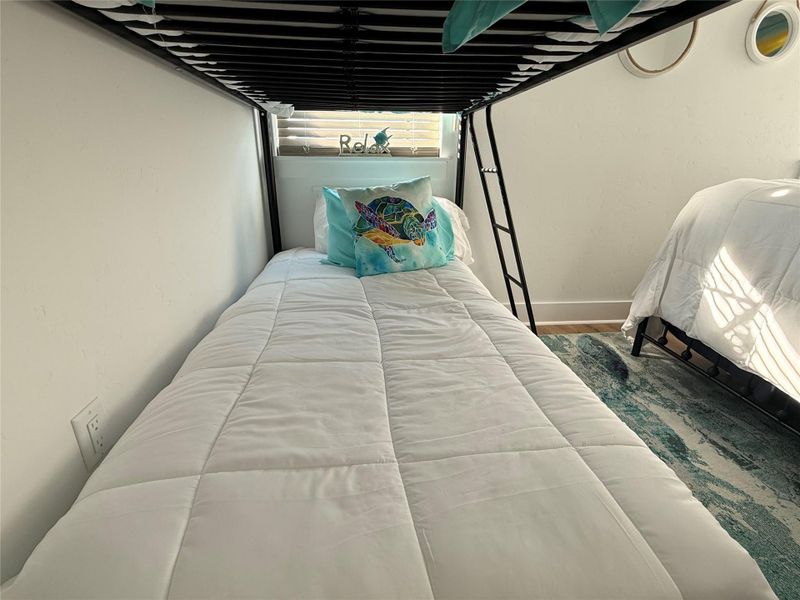 This cozy bedroom features a bunk bed setup with a colorful pillow, soft white bedding, and a nearby window for natural light. Perfect for a child’s room or guest space.