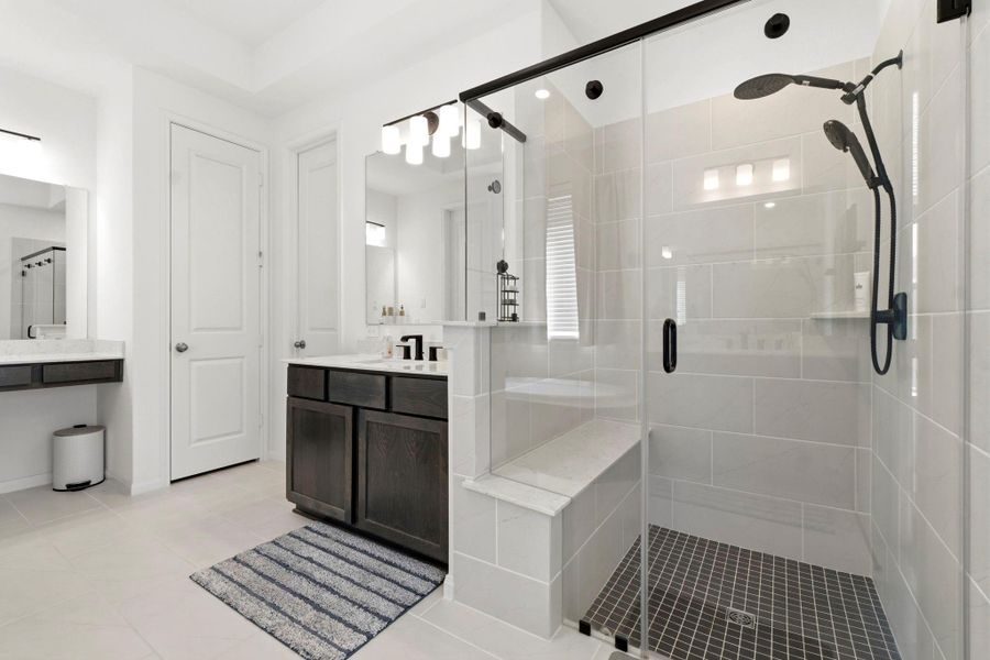 This modern bathroom features a spacious glass-enclosed shower with dual shower heads and a built-in bench. The room is bright, with sleek dark cabinetry contrasting against light walls and tiles, creating a clean and inviting atmosphere.