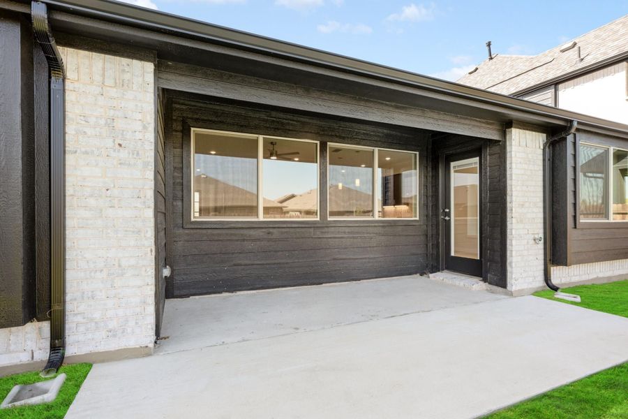 Covered back patio with dark wood siding, large windows, glass door, and concrete floor next to green lawn.