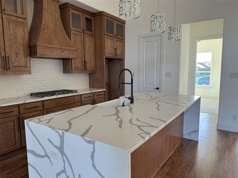 Kitchen featuring premium range hood, stainless steel gas cooktop, an island with sink, and pendant lighting