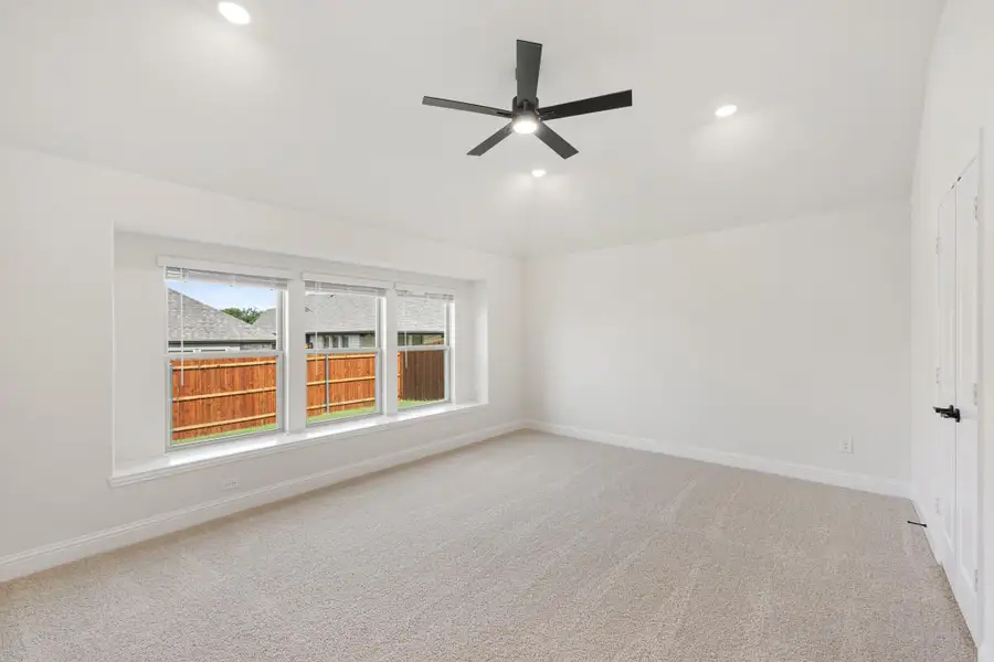 Empty bedroom with carpet flooring, black ceiling fan, and triple window overlooking backyard fence