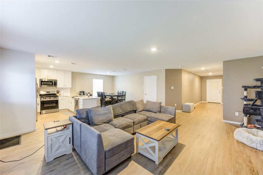This spacious open-concept living area features a modern kitchen with stainless steel appliances and white cabinetry, a cozy seating area with a sectional sofa, and wood-style flooring throughout. The room is well-lit with recessed lighting and has a neutral color palette, creating a warm and inviting atmosphere.