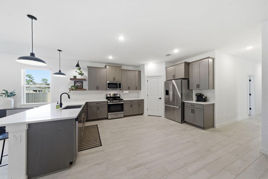 This modern kitchen features sleek gray cabinetry, stainless steel appliances, and a spacious layout with ample counter space. Natural light streams through a window, highlighting the stylish pendant lighting and open shelving for decor or storage.