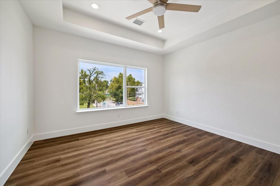 The spacious primary bedroom creates an airy retreat with warm wood-look flooring, crisp white walls, and a distinctive tray ceiling that adds architectural interest. A modern ceiling fan and recessed lighting enhance both comfort and style, while the oversized window fills the room with natural light and leafy neighborhood views. Generous dimensions provide ample room for a complete bedroom suite, sitting area, or additional furnishings while maintaining an open, relaxing atmosphere. The spacious primary bedroom creates an airy retreat with warm wood-look flooring, crisp white walls, and a distinctive tray ceiling that adds architectural interest. A modern ceiling fan and recessed lighting enhance both comfort and style, while the oversized window fills the room with natural light and leafy neighborhood views. Generous dimensions provide ample room for a complete bedroom suite, sitting area, or additional furnishings while maintaining an open, relaxing atmosphere.