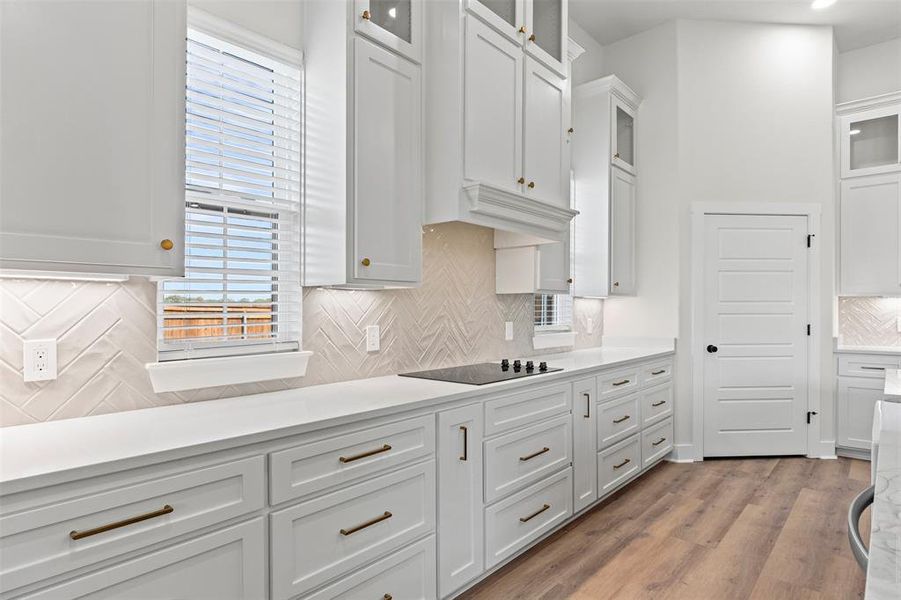 Kitchen with tasteful backsplash, glass insert cabinets, white cabinets, light wood finished floors, and light stone countertops Kitchen with tasteful backsplash, glass insert cabinets, white cabinets, light wood finished floors, and light stone countertops