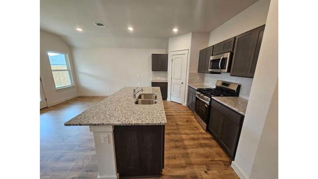Spacious kitchen featuring an elongated island with double sinks, dark wood-finish cabinetry, stainless steel appliances, granite countertops, and wood-finish flooring