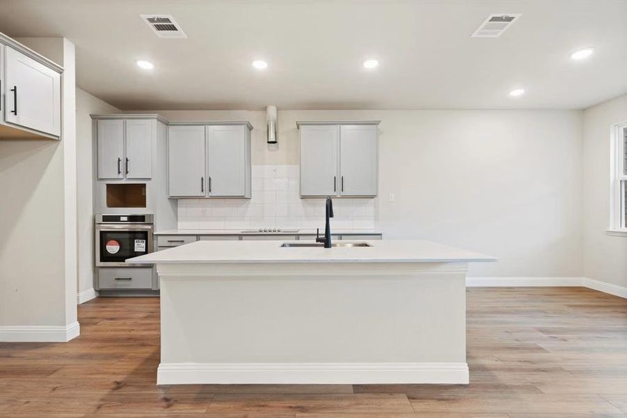 Kitchen featuring gray cabinets, sink, oven, light hardwood / wood-style floors, and a center island with sink