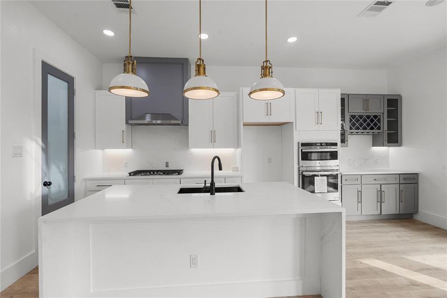 Kitchen with light stone countertops, light wood-style flooring, hanging light fixtures, stainless steel double oven, and a center island with sink Kitchen with light stone countertops, light wood-style flooring, hanging light fixtures, stainless steel double oven, and a center island with sink