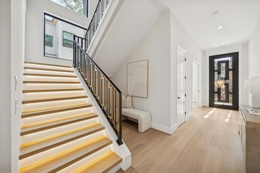 Guests are welcomed by a dramatic entry marked by a sleek glass-and-metal front door that opens to a spacious, sunlit corridor. The staircase itself is a design highlight, featuring illuminated stair treads that provide both ambiance and safety. A modern iron railing and expansive glass-pane windows with a lighted accent wall complete the dramatic architectural statement.