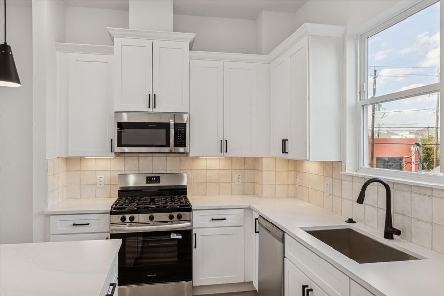 This kitchen features modern white cabinetry, stainless steel appliances including a gas stove and microwave, a sleek black faucet, and ample natural light from a large window. The clean design and neutral tile backsplash create a bright and inviting space. This kitchen features modern white cabinetry, stainless steel appliances including a gas stove and microwave, a sleek black faucet, and ample natural light from a large window. The clean design and neutral tile backsplash create a bright and inviting space.