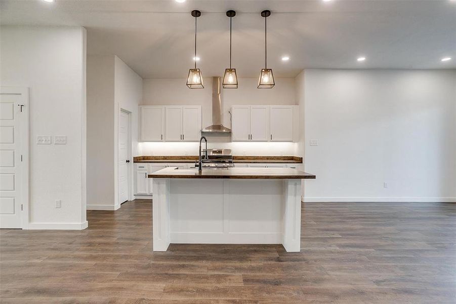 Kitchen with dark wood-type flooring, decorative light fixtures, a center island with sink, wall chimney range hood, and white cabinets