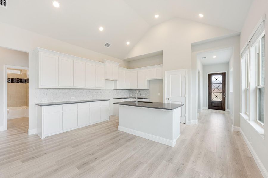 Open kitchen with white cabinets, dark countertop island, tile backsplash, and light hardwood floors under vaulted ceiling
