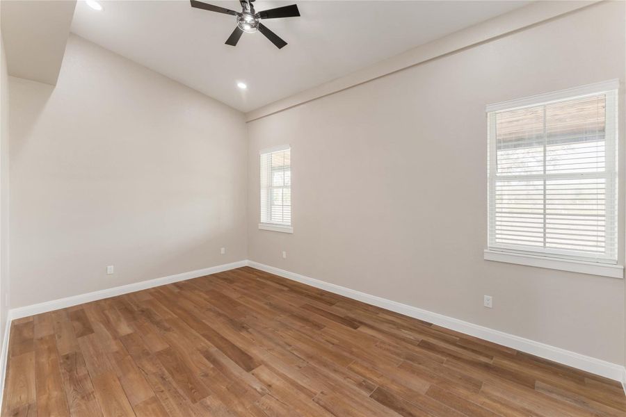 This room features a high ceiling with a modern ceiling fan, warm wood flooring, and neutral walls. It has large windows that allow natural light to fill the space, creating a bright and inviting atmosphere.