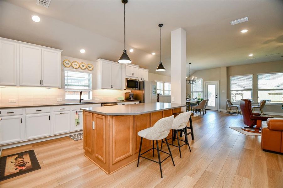 This photo showcases a spacious, open-concept kitchen and living area with light wood flooring. The kitchen features white cabinetry, modern appliances, and a large island with seating. Ample natural light flows through the windows, highlighting the cozy dining and living spaces.
