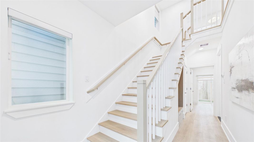 This photo showcases a bright, modern entryway with a wooden staircase featuring white railings. It has sleek hardwood floors, a large window, and a hallway leading to a glass door, creating an open and inviting atmosphere.