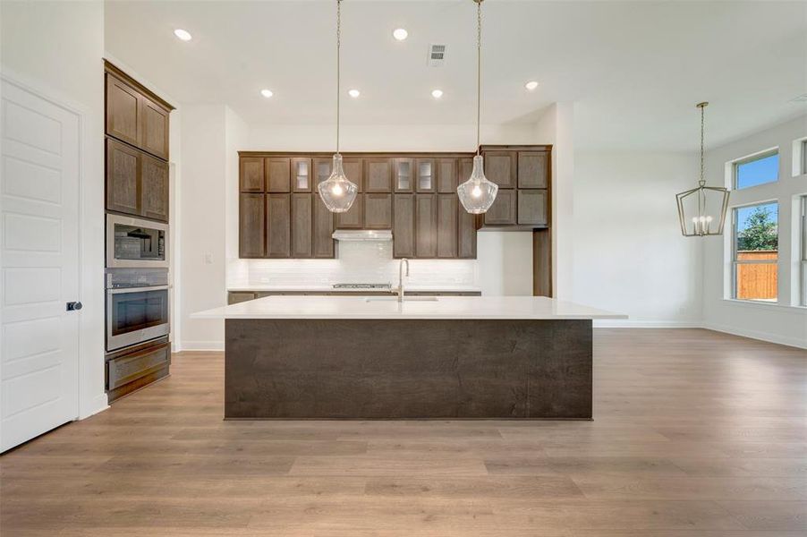 Modern kitchen featuring an expansive island with a light-toned countertop, wood-finish cabinetry, built-in stainless steel appliances, and wood-finish flooring Modern kitchen featuring an expansive island with a light-toned countertop, wood-finish cabinetry, built-in stainless steel appliances, and wood-finish flooring
