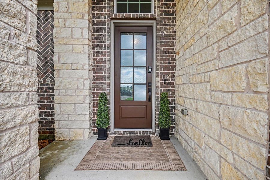 This photo showcases a welcoming front entrance with a brown door featuring glass panels, framed by brick and stone walls. Flanking the door are two potted plants, and a doormat with "hello" adds a friendly touch. This photo showcases a welcoming front entrance with a brown door featuring glass panels, framed by brick and stone walls. Flanking the door are two potted plants, and a doormat with "hello" adds a friendly touch.