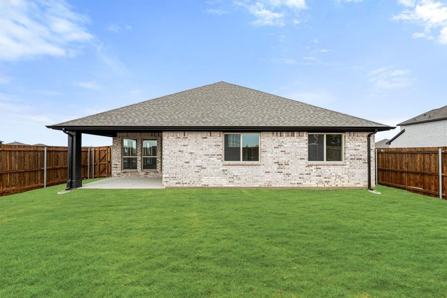 Rear view of single-story brick home with covered patio, large green lawn, and wood privacy fence