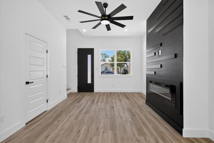 Spacious room featuring wood-finish flooring, a contemporary black accent wall with an integrated linear fireplace, and a stylish ceiling fan Spacious room featuring wood-finish flooring, a contemporary black accent wall with an integrated linear fireplace, and a stylish ceiling fan