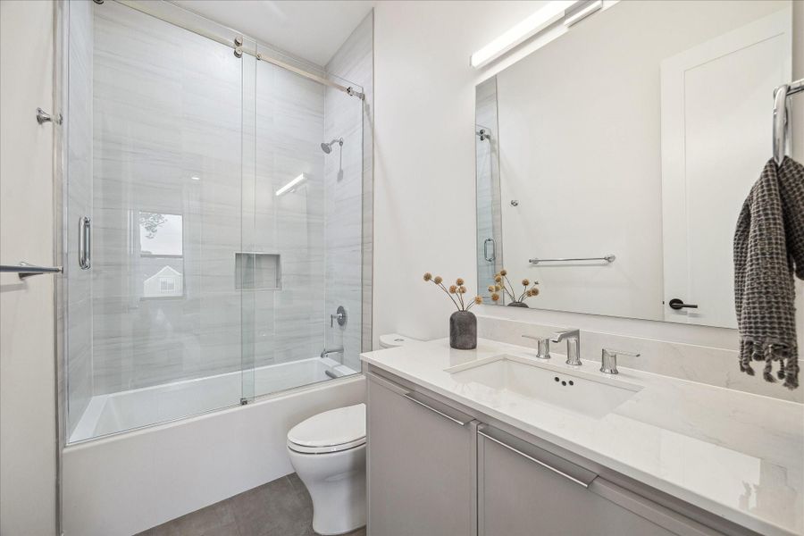 Bright secondary bath featuring a sleek vanity, expansive mirror, and a glass-enclosed tub and shower combination with modern tile surround. Clean lines, neutral tones, and polished fixtures create a fresh, contemporary space designed for comfort and everyday functionality.