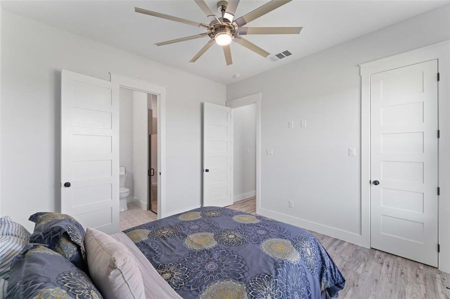 Bedroom featuring an overhead ceiling fan with lighting, wood-finish flooring, white paneled doors with black hardware, and an adjacent bathroom with a toilet and shower enclosure
