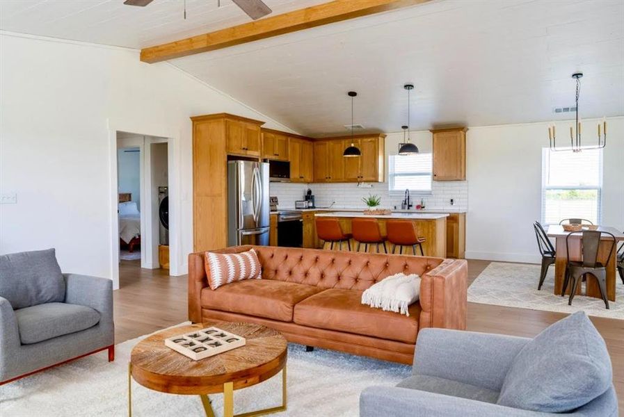 The open concept living area features light wood flooring, a white ceiling with exposed wood beams, and a modern kitchen with a white subway tile backsplash, light wood cabinetry, and stainless steel appliances