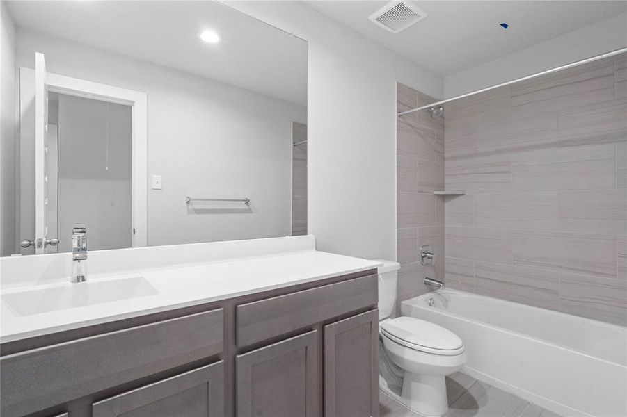 Secondary bath features tile flooring, bath/shower combo with tile surround, stained wood cabinets, beautiful light countertops, mirror, dark, sleek fixtures and modern finishes.