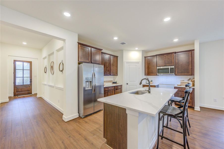 This kitchen features a modern design with stainless steel appliances, a large island with seating, dark wood cabinets, and recessed lighting. The open layout flows into a bright, inviting entryway with hardwood flooring.