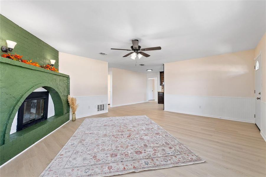 Unfurnished living room featuring light wood finished floors, a wainscoted wall, a brick fireplace, and ceiling fan