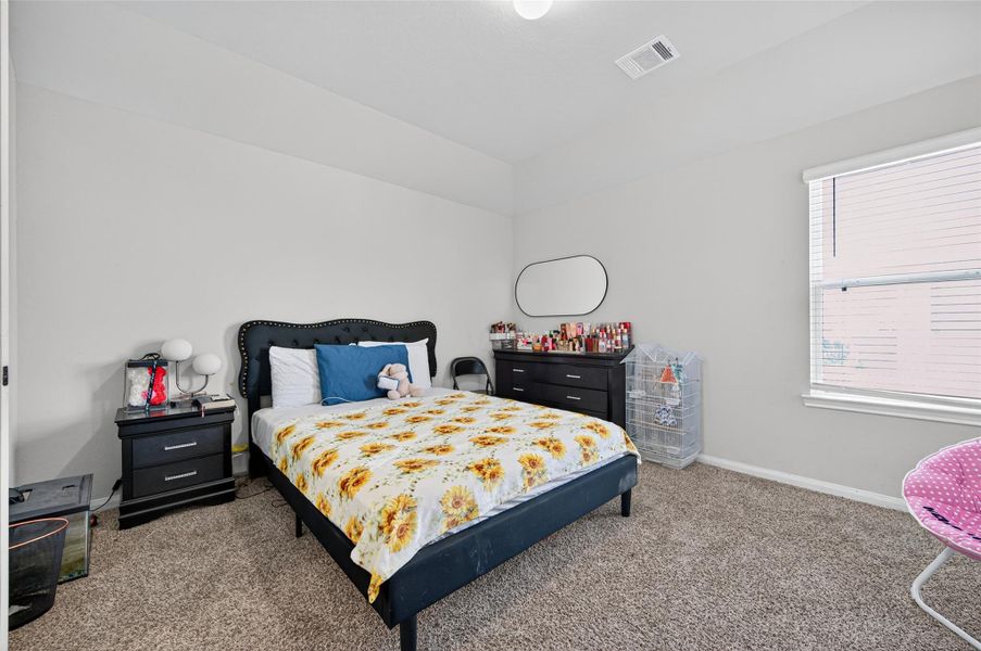 This cozy bedroom features neutral walls and carpet, a large window for natural light, and is furnished with a bed, nightstand, and dresser. The space is inviting and well-suited for relaxation.
