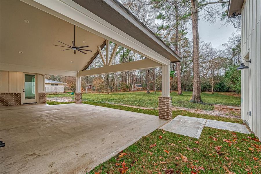 covered patio with a ceiling fan, offering a view of a large, backyard bordered by mature trees with walkway to the shop