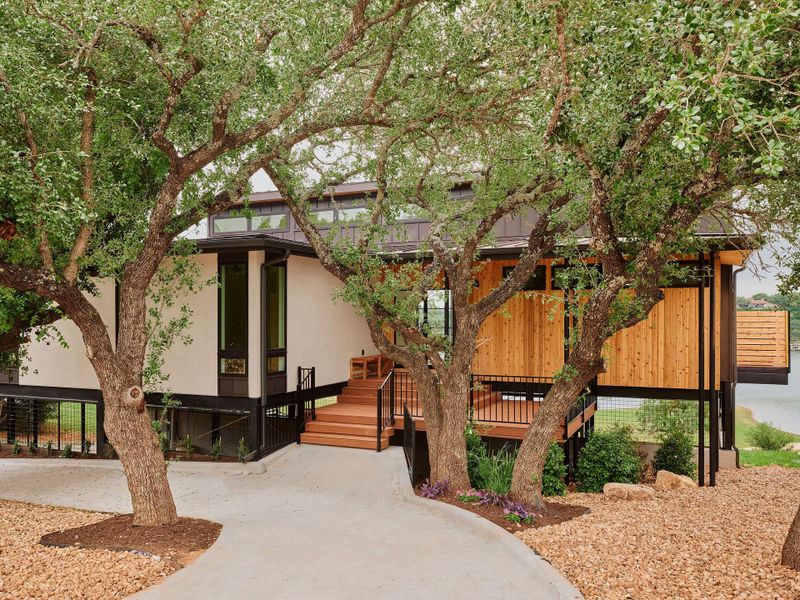 Contemporary architecture featuring a white and natural wood-paneled exterior, multiple mature trees, a concrete walkway, and a prominent deck overlooking a body of water
