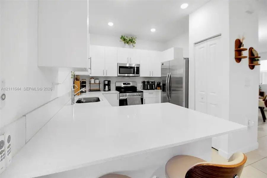 Kitchen featuring white shaker-style cabinetry, a solid-surface breakfast bar, stainless steel appliances, and recessed lighting Kitchen featuring white shaker-style cabinetry, a solid-surface breakfast bar, stainless steel appliances, and recessed lighting