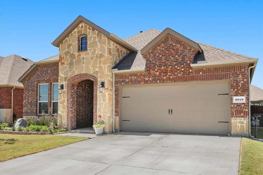 French country inspired facade featuring stone siding, a shingled roof, brick siding, and driveway French country inspired facade featuring stone siding, a shingled roof, brick siding, and driveway