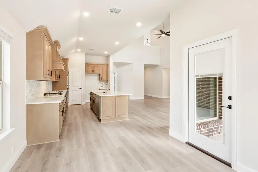 Open kitchen with tan cabinets, center island, light wood floors, and vaulted ceiling opening to living area Open kitchen with tan cabinets, center island, light wood floors, and vaulted ceiling opening to living area