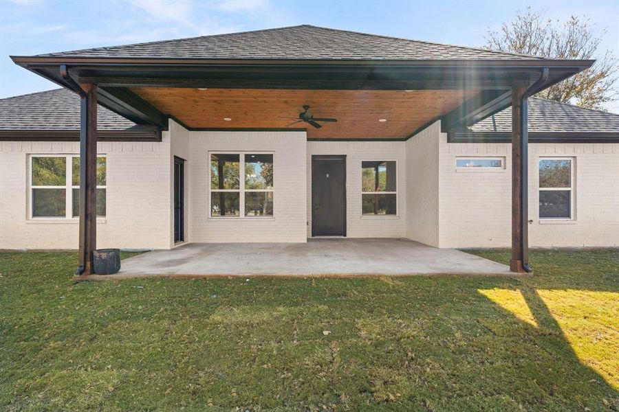 Back of property with a ceiling fan, brick siding, a patio area, a yard, and roof with shingles