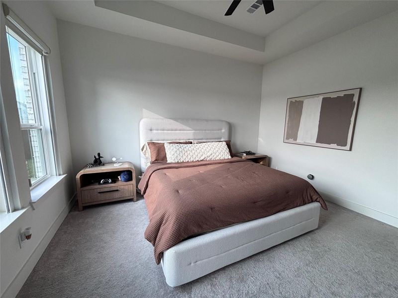The master bedroom is spacious and bright, with a contemporary design. It has a ceiling fan, multiple windows offering natural light and a view, and is complemented by neutral tones and modern furnishings
