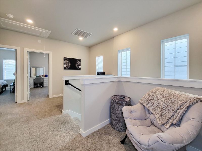 Carpeted landing area on an upper floor, featuring soft lighting and neutral walls. It includes a comfortable chair with a blanket, a decorative basket, and two doorways leading to adjacent rooms. The space is brightened by windows, creating a welcoming atmosphere.