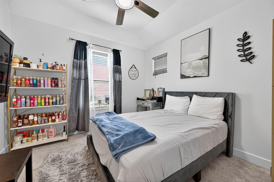 This cozy bedroom features a comfortable bed, soft carpeting, and ample natural light from two windows. It includes a ceiling fan, stylish wall decor, and shelving for storage or display. This cozy bedroom features a comfortable bed, soft carpeting, and ample natural light from two windows. It includes a ceiling fan, stylish wall decor, and shelving for storage or display.