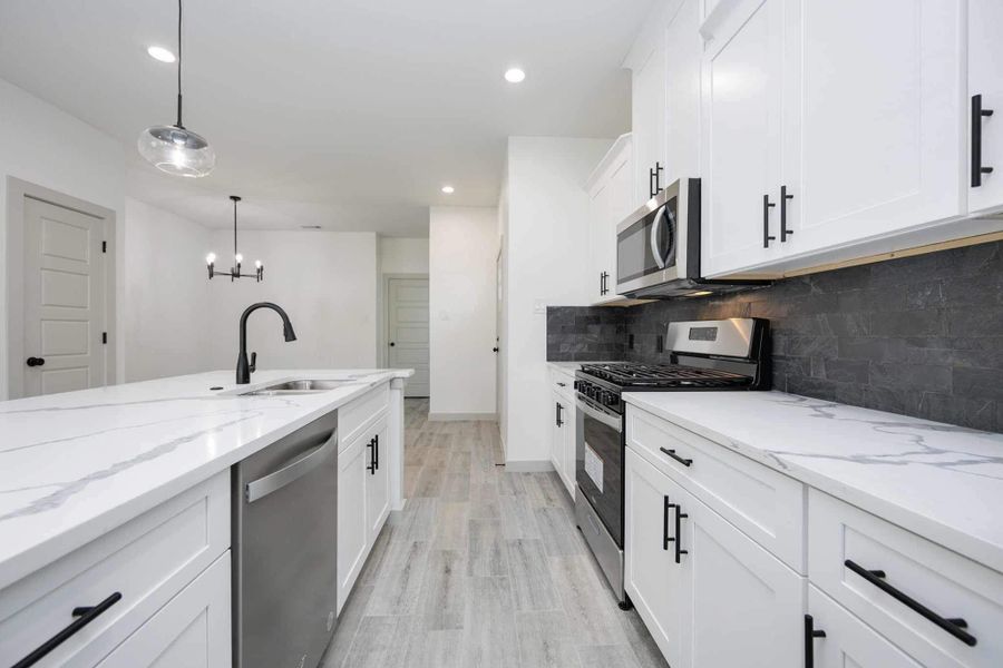 Bright and open kitchen featuring quartz countertops, shaker-style cabinetry, stainless steel appliances, and a striking dark tile backsplash. The oversized island offers additional prep space, seating potential, and modern pendant lighting above.