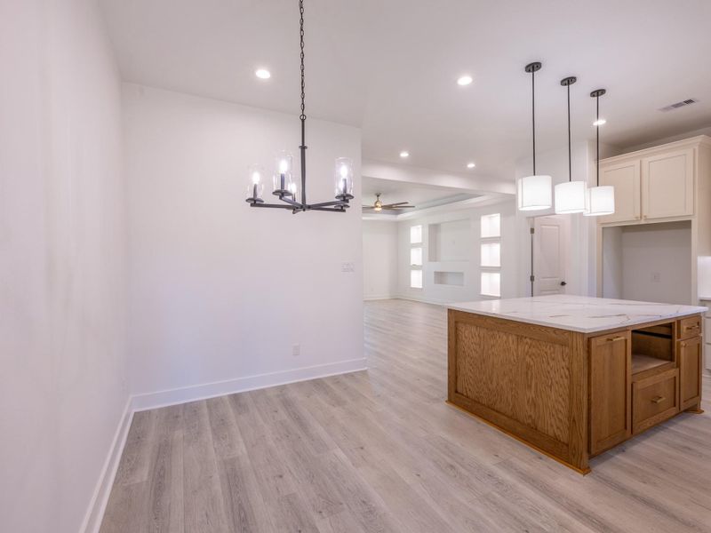 This photo showcases a bright, open-concept living area with light wood flooring. It features a modern kitchen island with pendant lighting and a nearby dining space with a stylish chandelier. The area flows into a living room with a ceiling fan and built-in wall shelving.