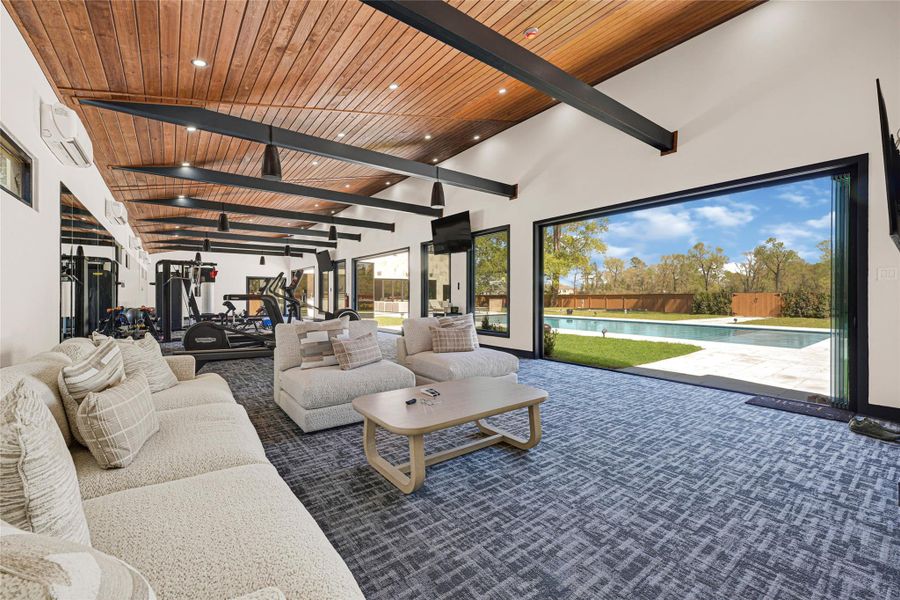Gym features a stylish wood-paneled ceiling with exposed beams, modern exercise equipment, and a cozy lounge area. Large sliding doors open to a beautiful patio and pool, offering a seamless indoor-outdoor experience.