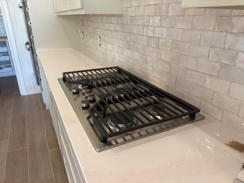 Kitchen view of stainless steel gas stovetop, decorative backsplash, light stone countertops, and white cabinets