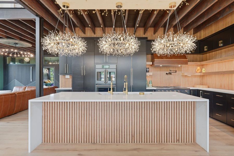 This modern kitchen features a spacious island with a sleek white countertop and wood accents. Elegant, contemporary chandeliers add a touch of luxury, while dark cabinets provide ample storage. The open layout connects seamlessly to a stylish living area.