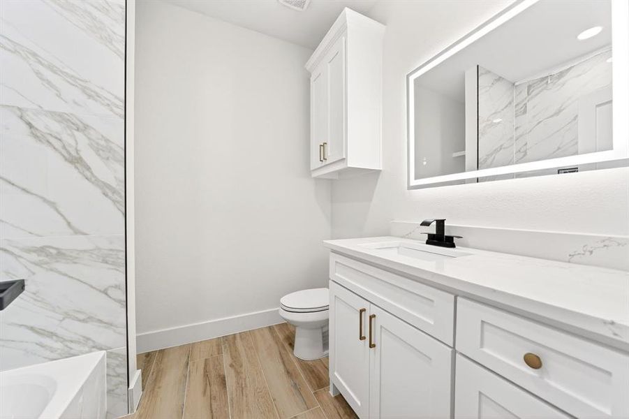 Vanity area featuring an illuminated mirror, white shaker-style cabinetry with gold-tone hardware, and a white countertop with integrated sink Vanity area featuring an illuminated mirror, white shaker-style cabinetry with gold-tone hardware, and a white countertop with integrated sink