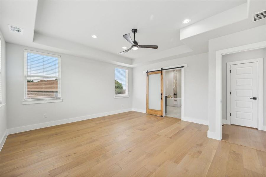 Unfurnished bedroom with a barn door, ceiling fan, a raised ceiling, and light hardwood / wood-style flooring