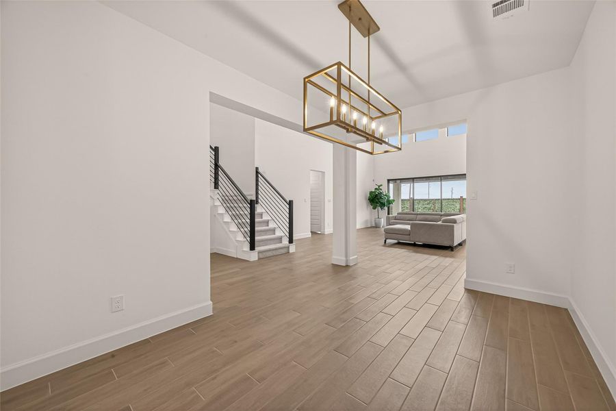 This photo showcases a modern, open-concept living space with sleek wood flooring, a contemporary chandelier, and a stylish staircase with black railing. The room is bright and inviting, featuring high ceilings and large windows that offer plenty of natural light.